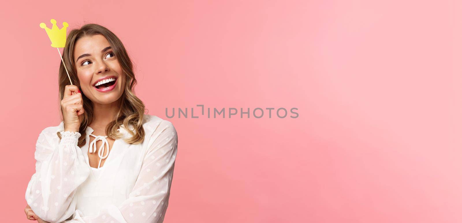 Spring, happiness and celebration concept. Close-up portrait of dreamy beautiful young blond girl imaging something cute and romantic, smiling look up daydreaming with crown on stick, pink background by Benzoix