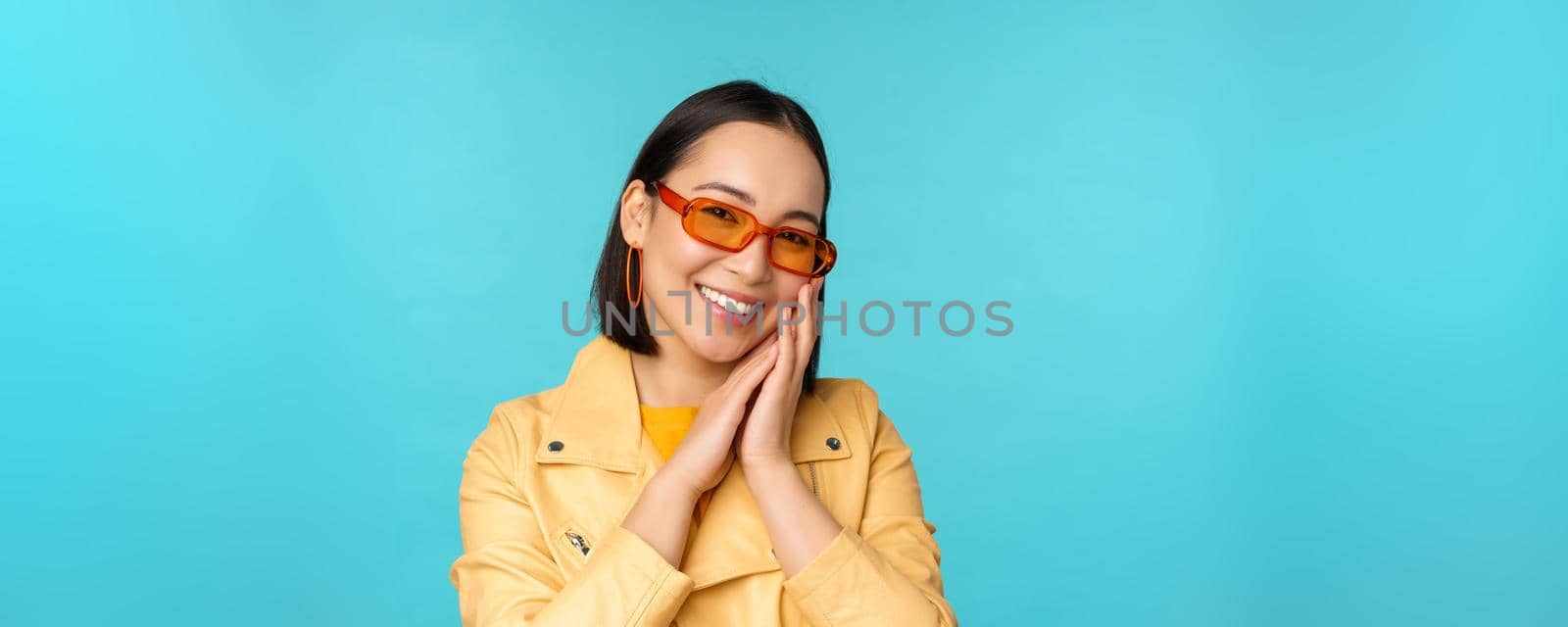 Close up portrait of asian young woman in sunglasses, smiling and looking romantic, standing happy over blue background. Copy space