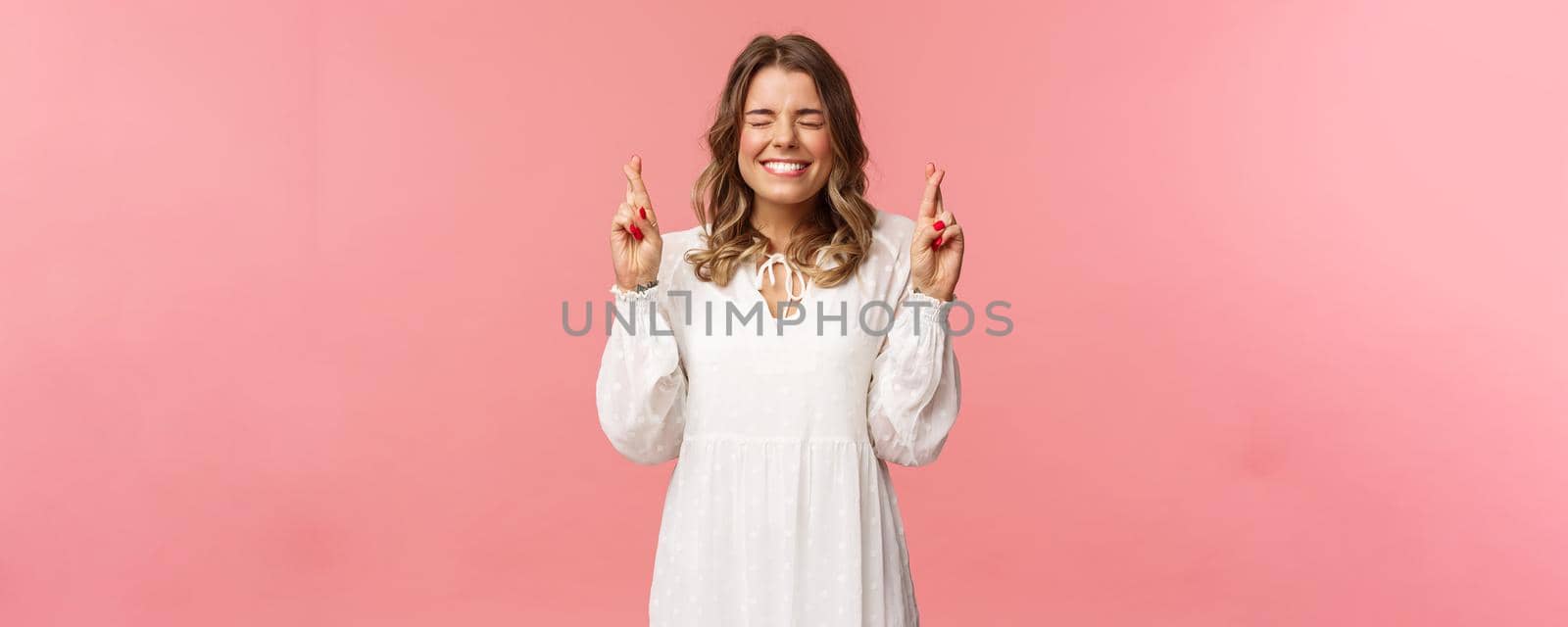 Portrait of hopeful lucky cute blond girl in white dress praying, anticiapte miracle, making wish to win, cross fingers good luck, smiling and close eyes and pleading god, pink background.