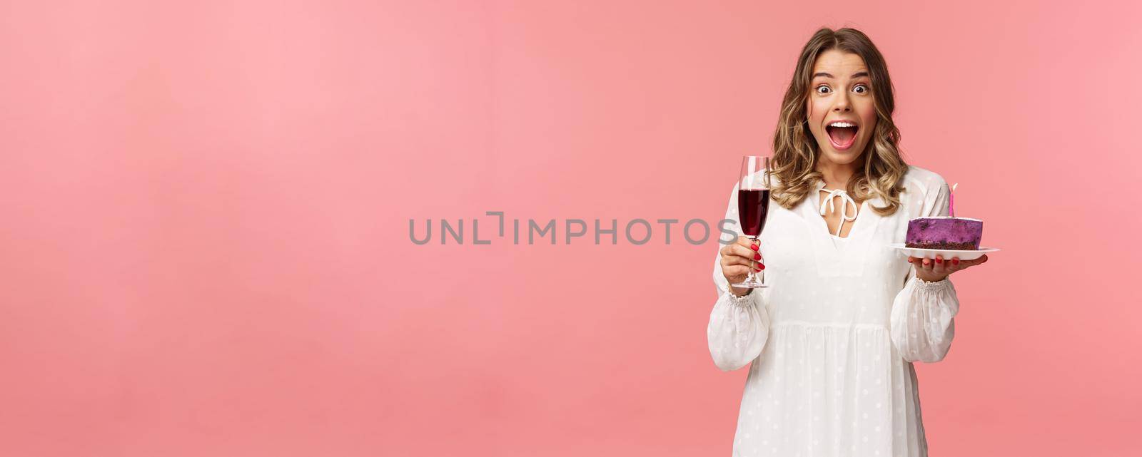 Holidays, spring and party concept. Portrait of fascinated and amused blond girl in white dress look astonished camera, celebrating birthday with b-day cake and glass of wine, pink background.