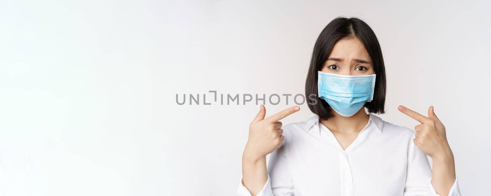 Close up of sad asian girl in medical face mask pointing at her head and looking upset, standing over white background.