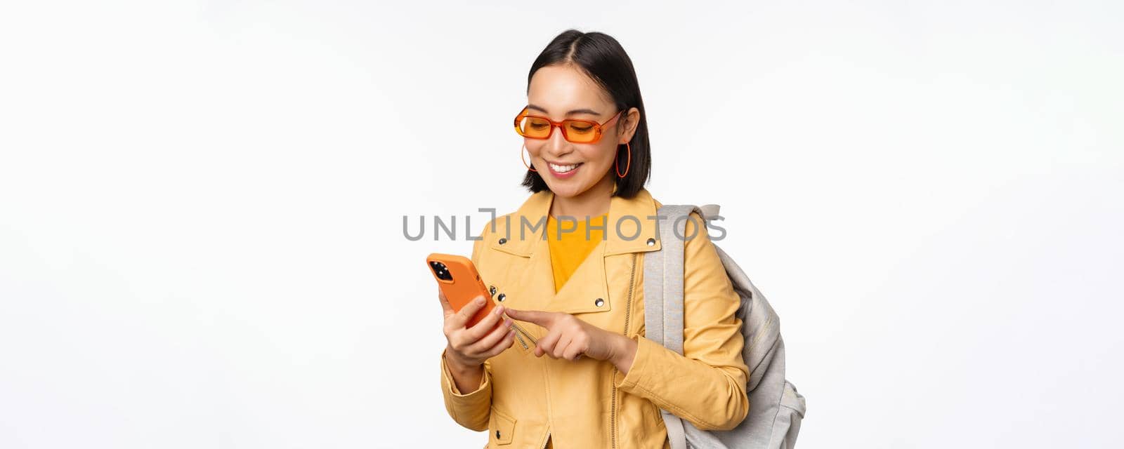 Stylish young asian woman tourist, traveller with backpack and smartphone smiling at camera, posing against white background. Copy space