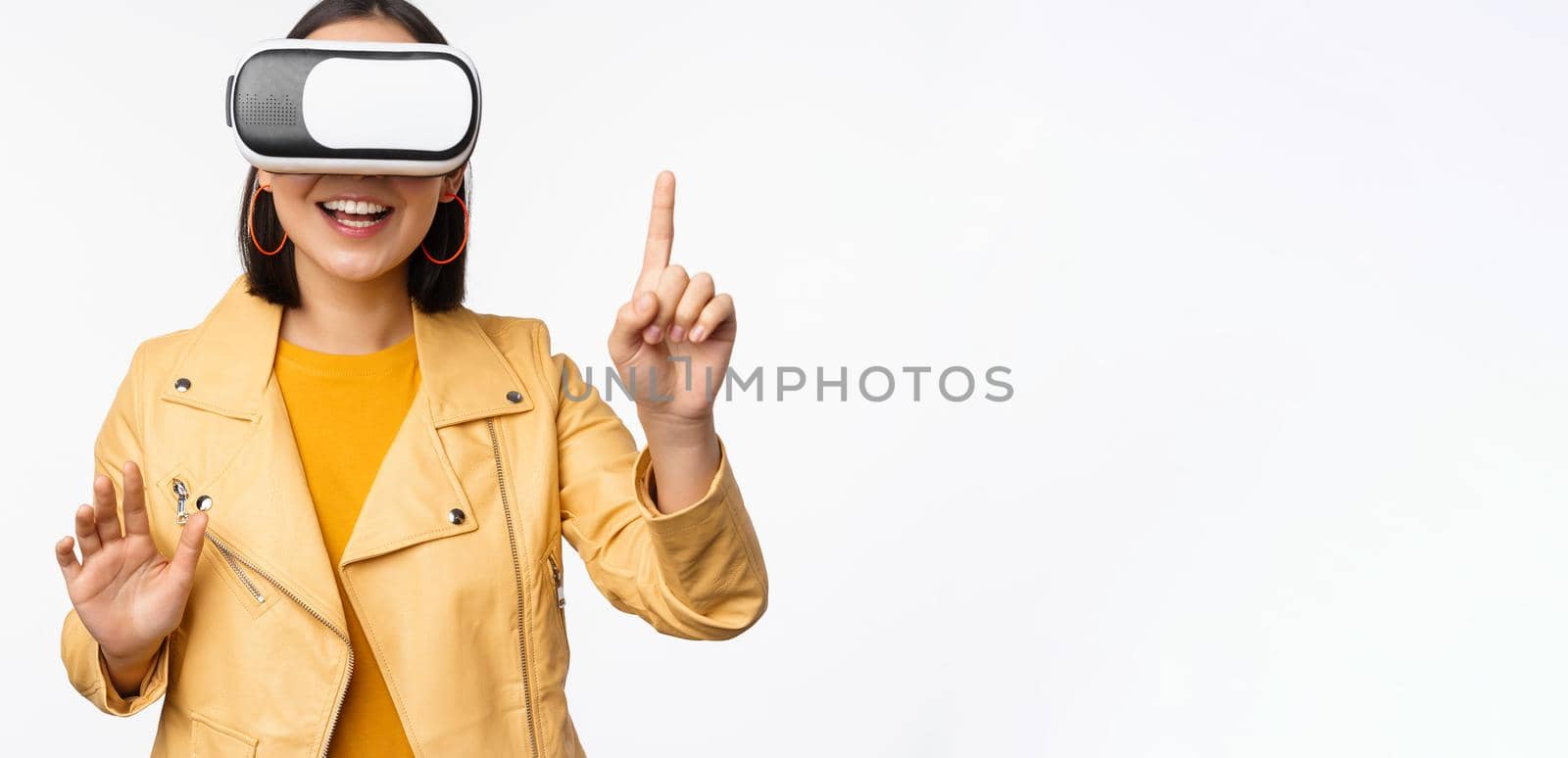Image of young asian woman wearng VR headset, using virtual reality glasses and smiling, pointing at smth, choosing smth, standing over white background.