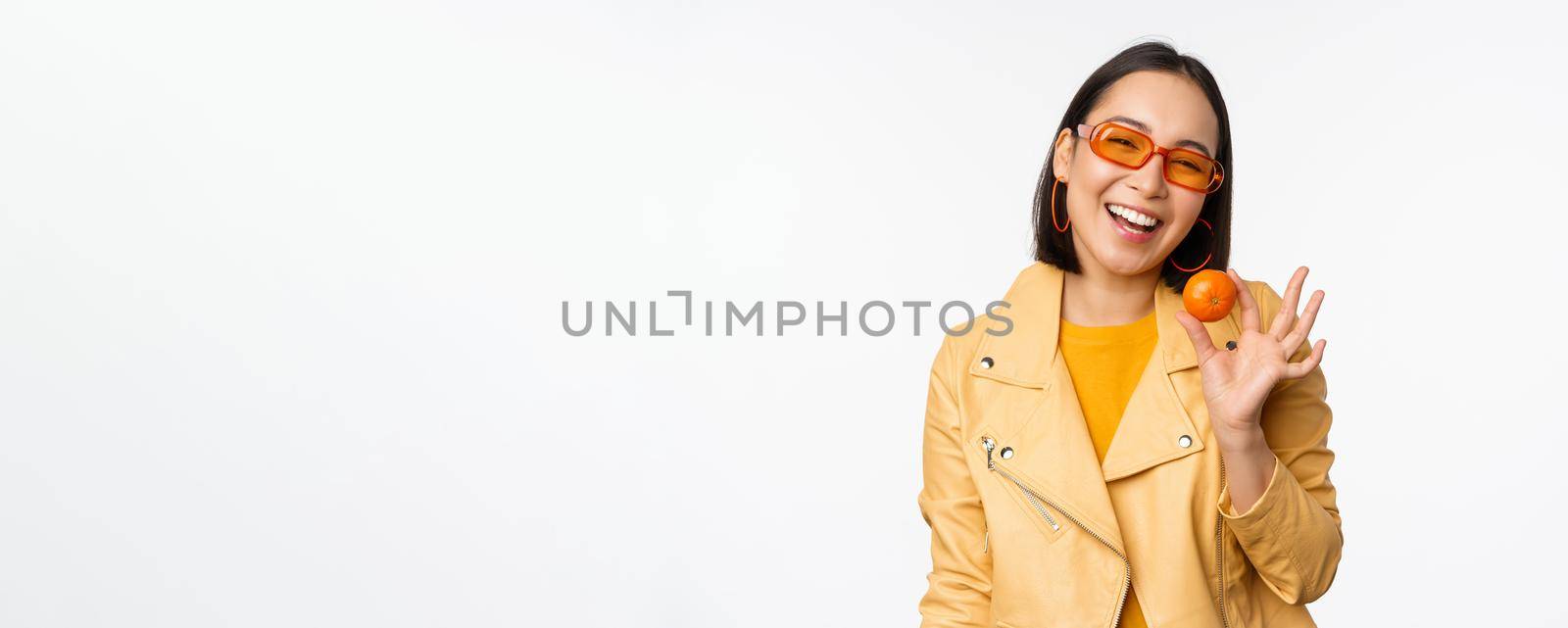 Beautiful asian girl in sunglasses showing tangerine and smiling, looking happy, posing in yellow against studio background.