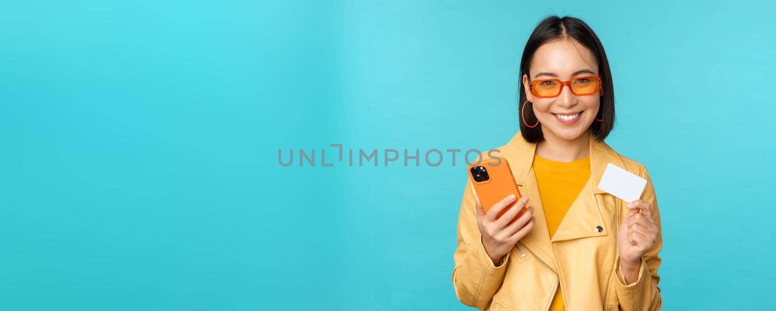 Online shopping. Stylish young asian woman in sunglasses, showing credit card and using smartphone, paying in internet, making purchase, standing over blue background.