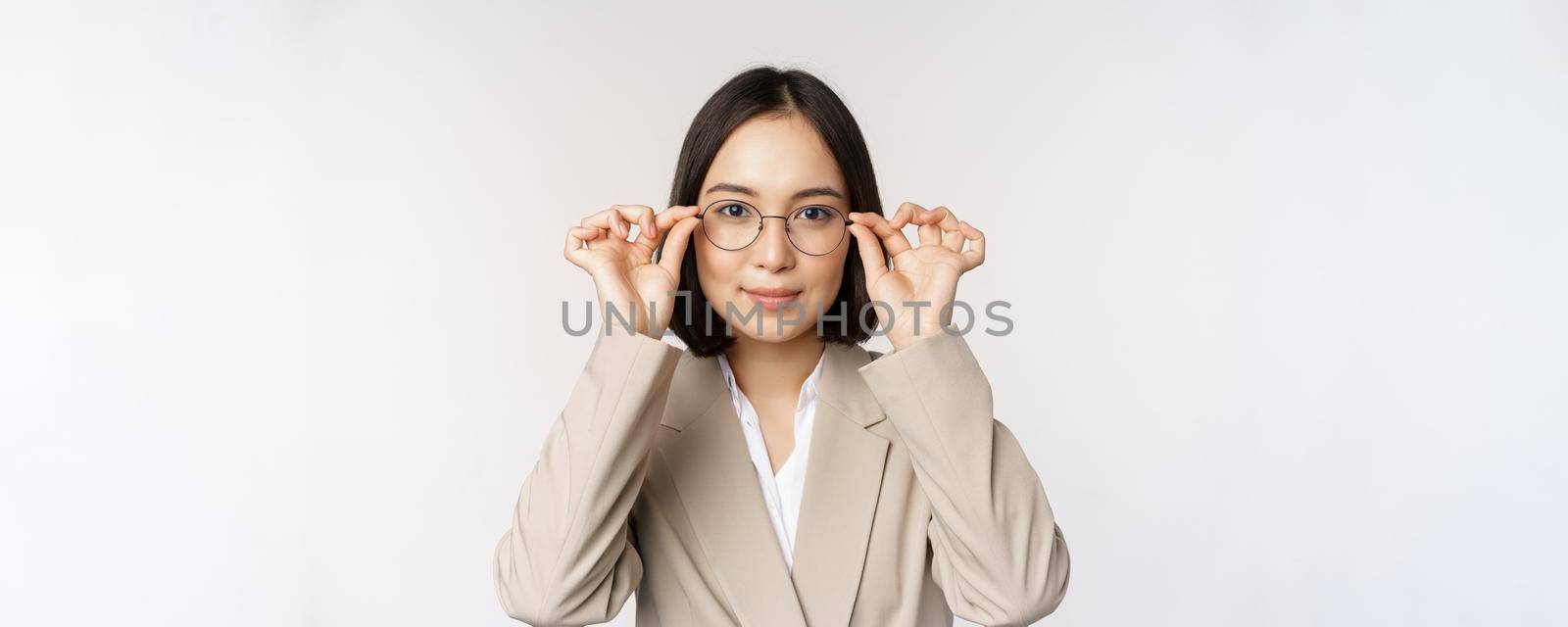 Smiling asian businesswoman trying new glasses, wearing eyewear, standing in suit over white background. Copy space