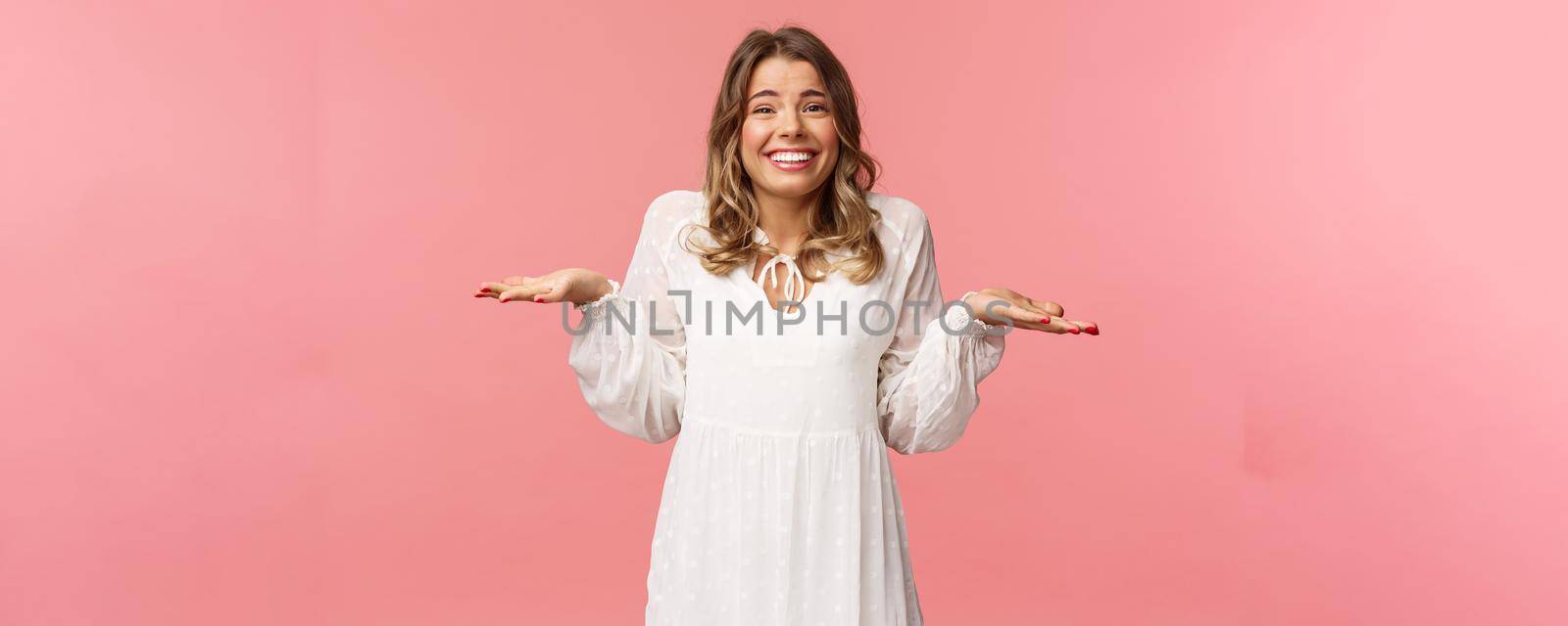 Beauty, fashion and women concept. Sorry dont know, cant say. Cheerful cute silly blond girl 20s, wear white dress, shrugging with hands sideways make awkward smile, apologizing, pink background.