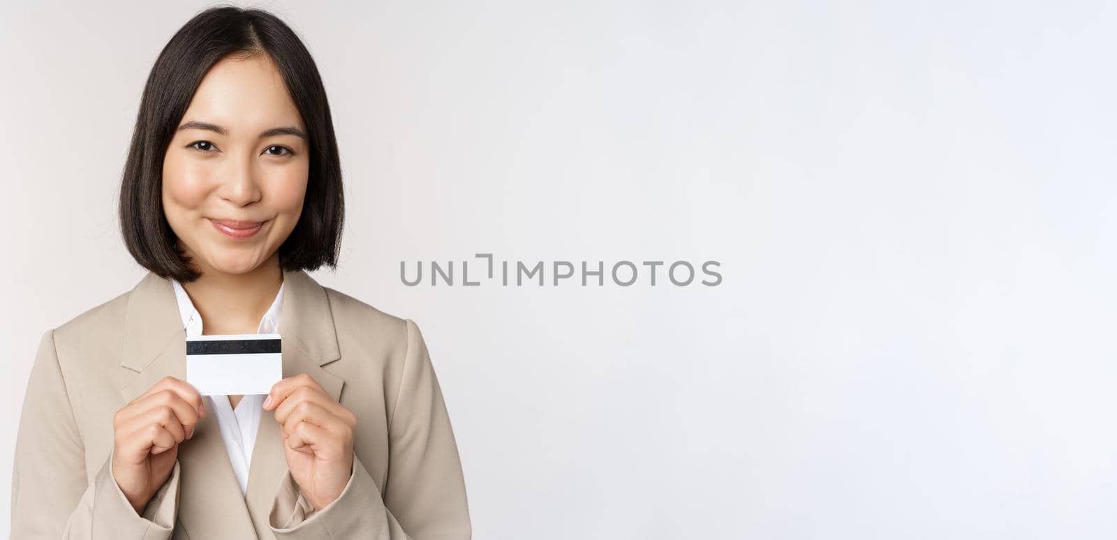 Smiling office clerk, asian corporate woman showing credit card, standing over white background in beige suit. Copy space