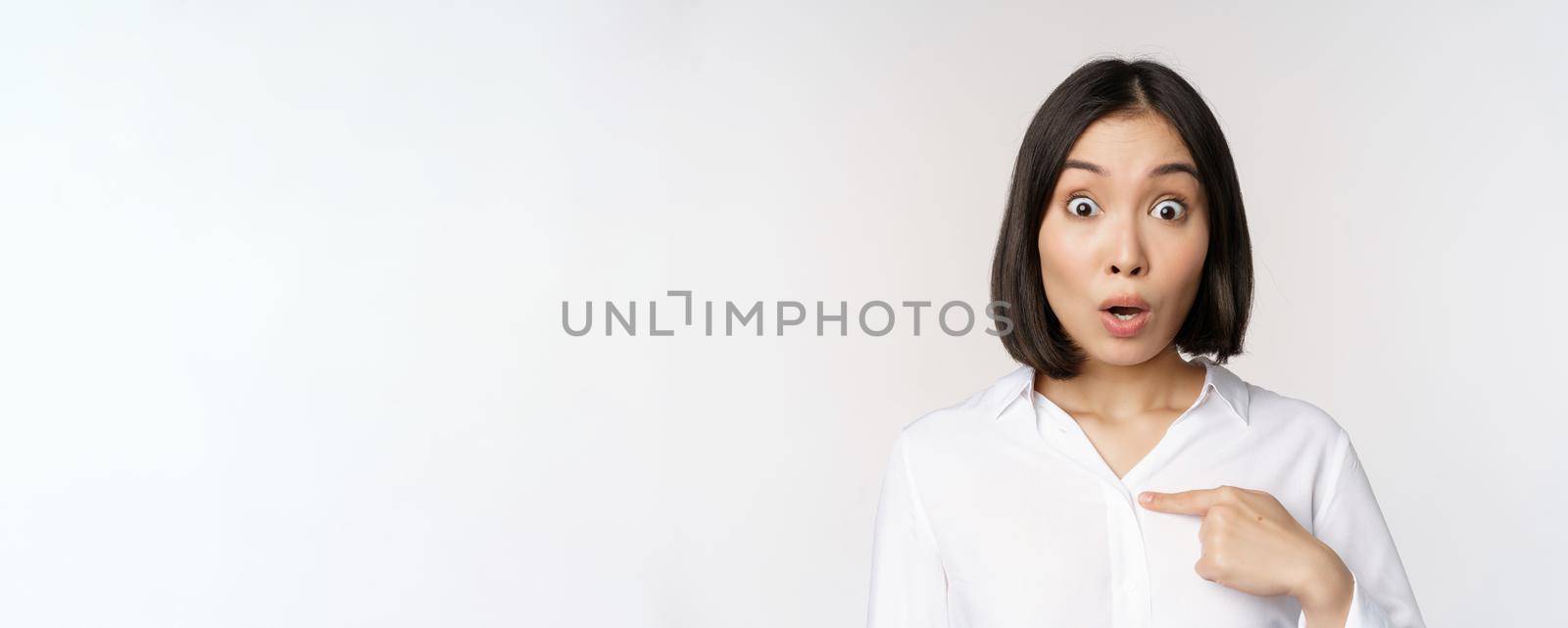 Close up portrait of young asian woman pointing finger at herself with disbelief, surprised that she was named, standing over white background.