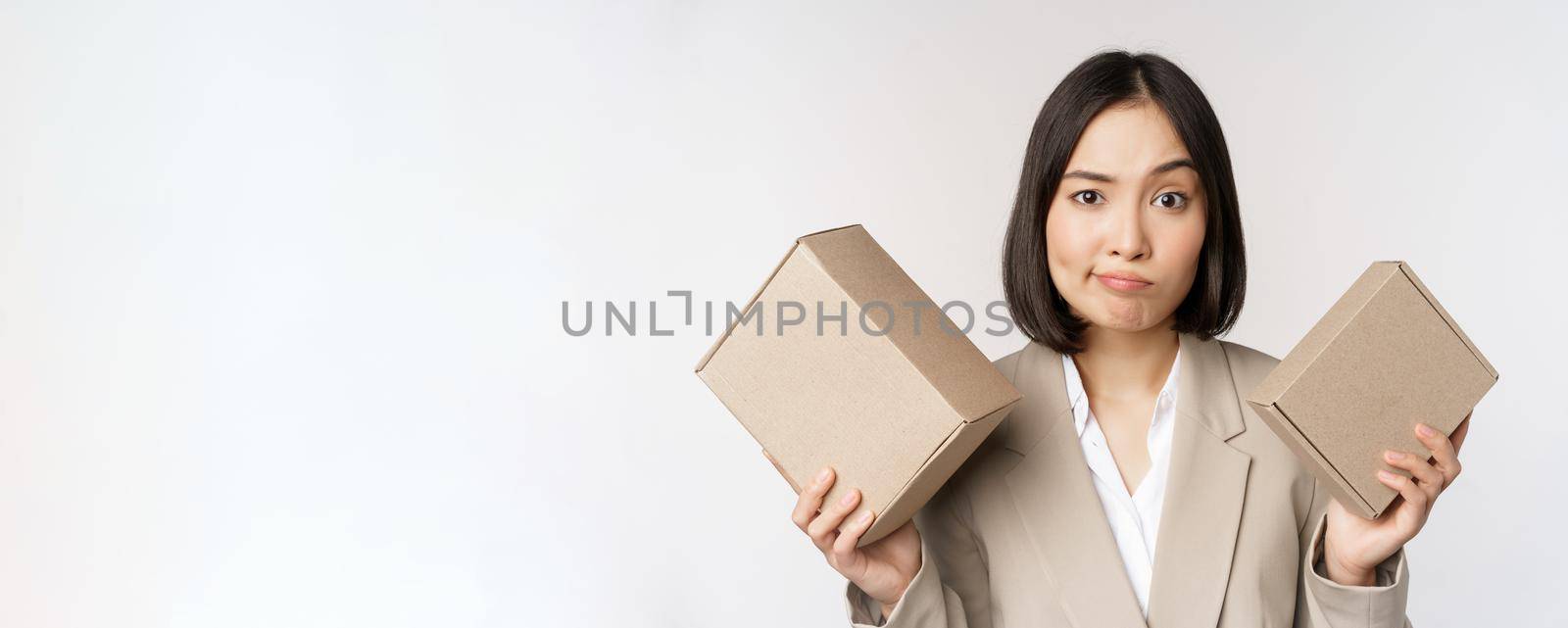 Puzzled asian saleswoman holding boxes, looking confused, standing in business suit over white background. Copy space