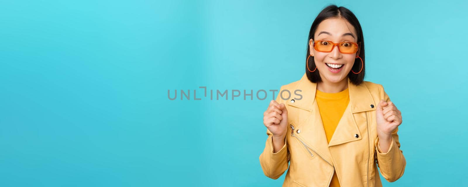 Image of enthusiastic young asian woman celebrating, triumphing, looking surprised and happy, clapping hands satisfied, standing over blue background.