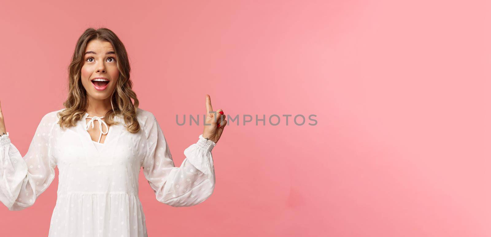 Waist-up portrait of beautiful young and healthy blond girl with cheerful, excited expression looking and pointing up, holding breath from excitement seeing something wonderful, pink background by Benzoix