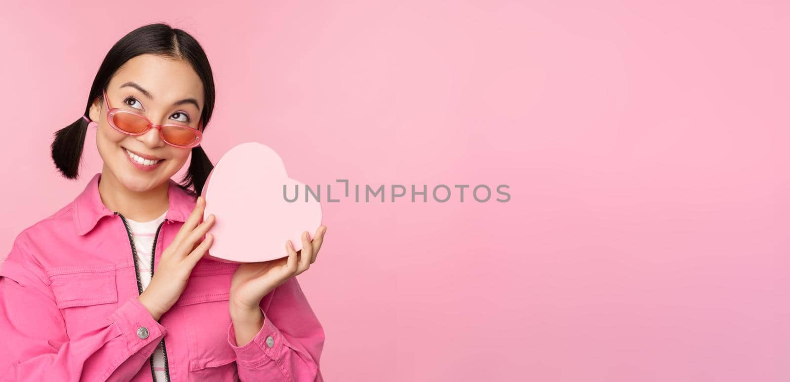 Image of stylish asian girlfriend in sunglasses, guessing whats inside gift box, heart-shaped present, standing over pink background.