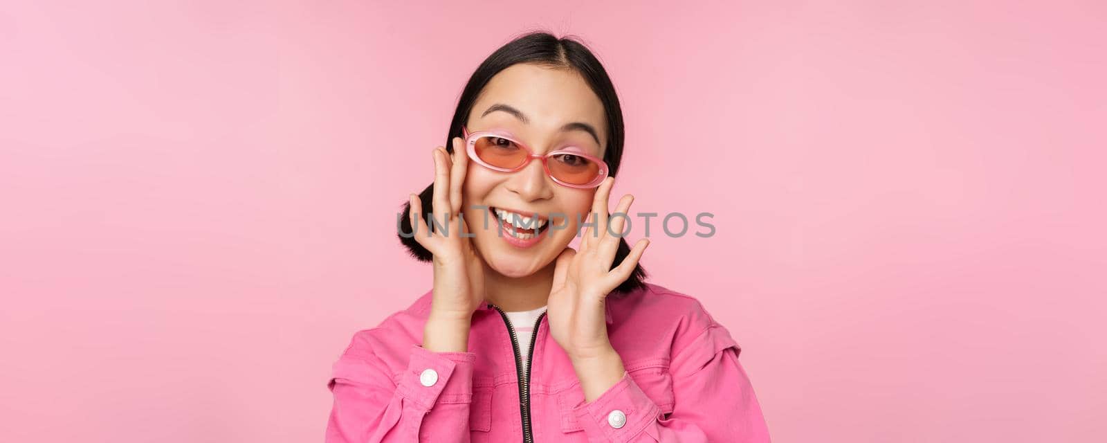 Cute modern japanese girl in sunglasses, smiling and looking happy, posing against pink background in stylish clothing.