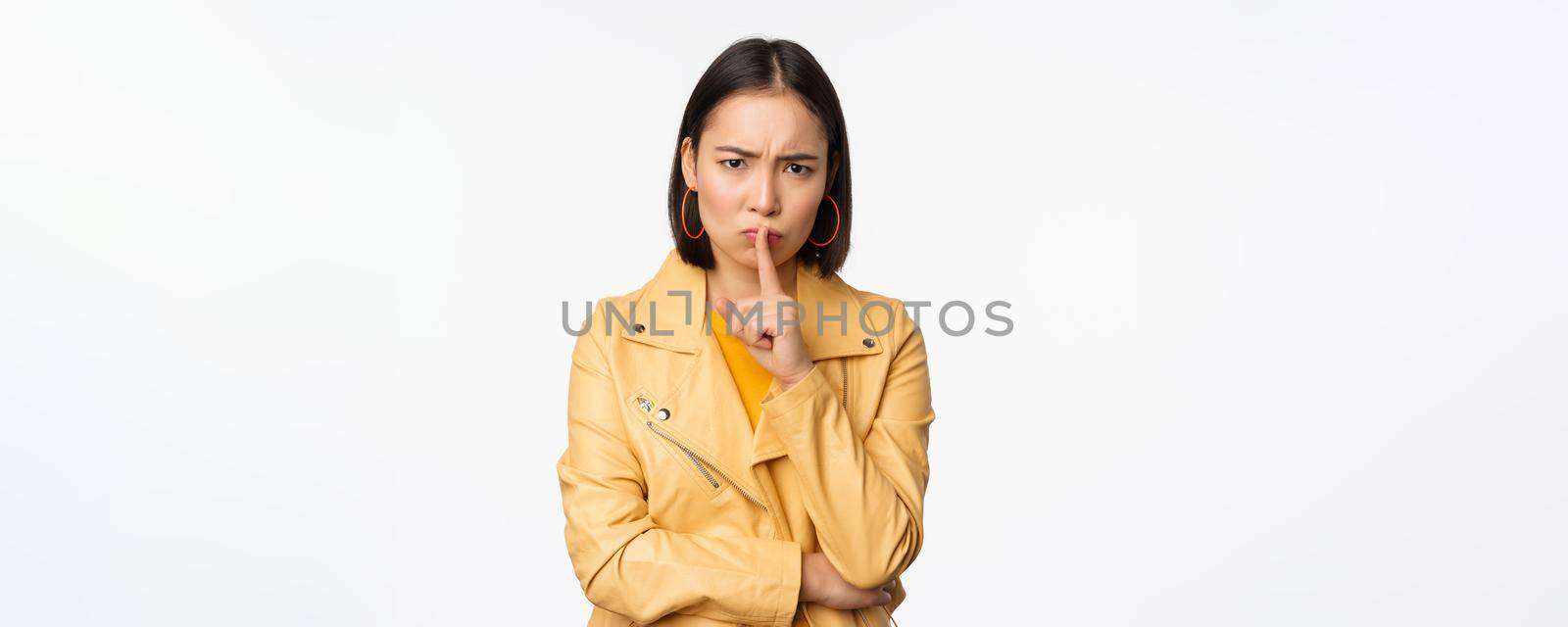 Portrait of angry korean girl shushing, woman frowning and tell to be quiet, showing hush, shh gesture, press finger to lips, taboo sign, standing over white background.