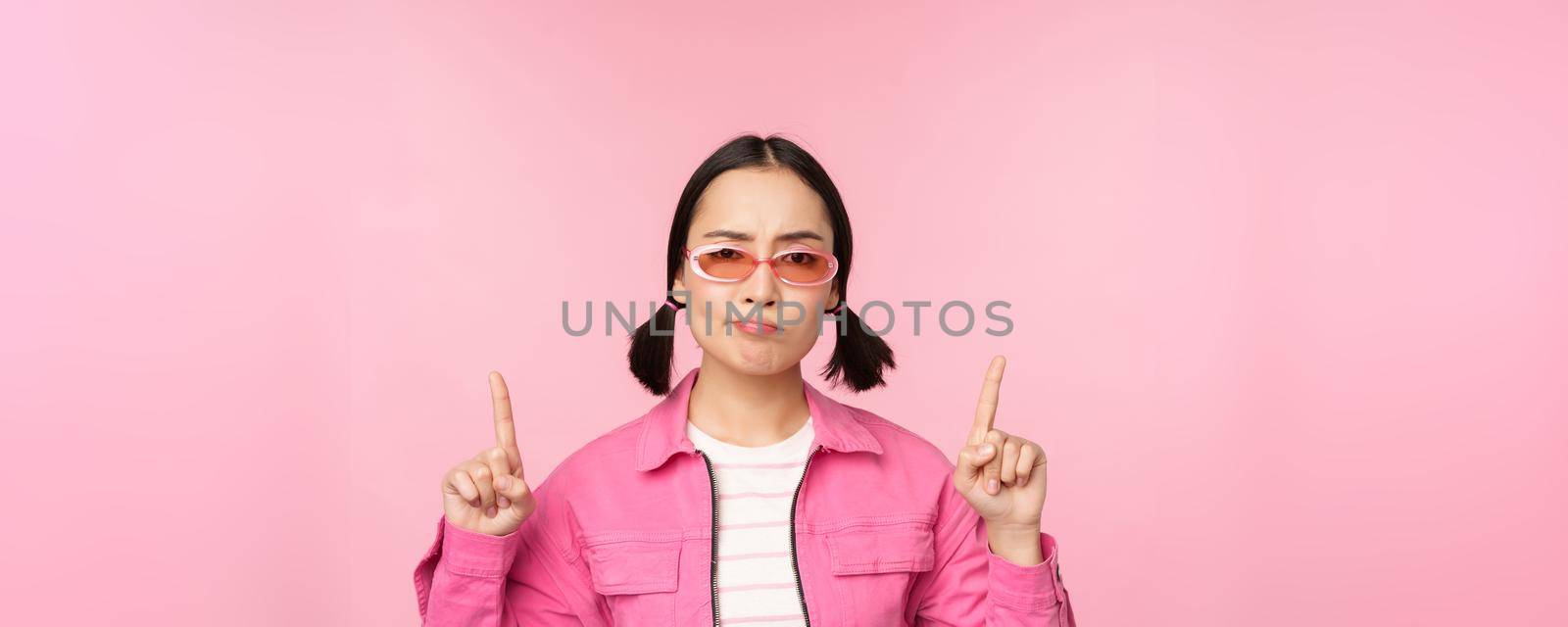 Close up of stylish korean girl, looking perplexed, pointing fingers up, showing advertisement, standing over pink background.