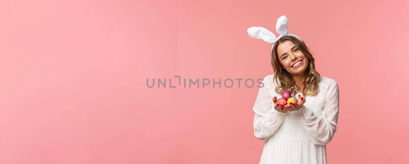 Holidays, spring and party concept. Portrait tender, romantic blond girl in white dress and rabbit ears, tilt head cute, smiling happy as holding painted eggs, celebrating Easter, pink background by Benzoix