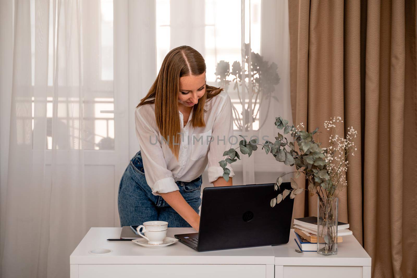 European professional woman is sitting with a laptop at a table in a home office, a positive woman is studying while working on a PC. She is wearing a beige jacket and jeans and is on the phone. by Matiunina