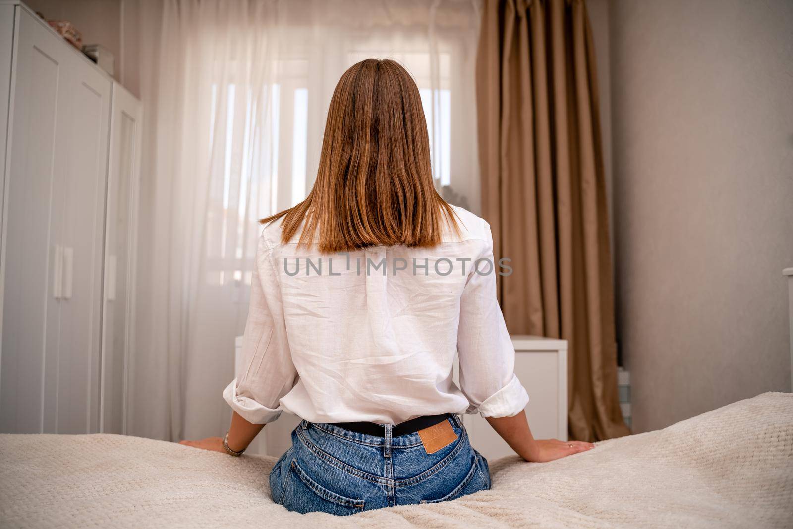 A young woman in a white shirt and jeans sits on the bed with her back against the background of the room and the window. Home furnishings