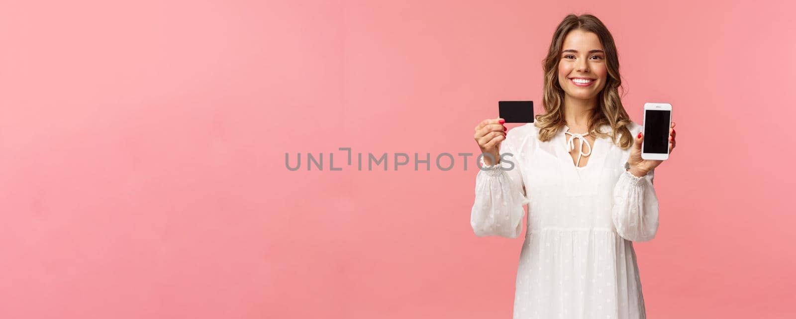 Portrait of satisfied cute blond girl in white dress, showing you her credit card and mobile phone application, online shopping app or bank website, smiling camera, standing pink background.