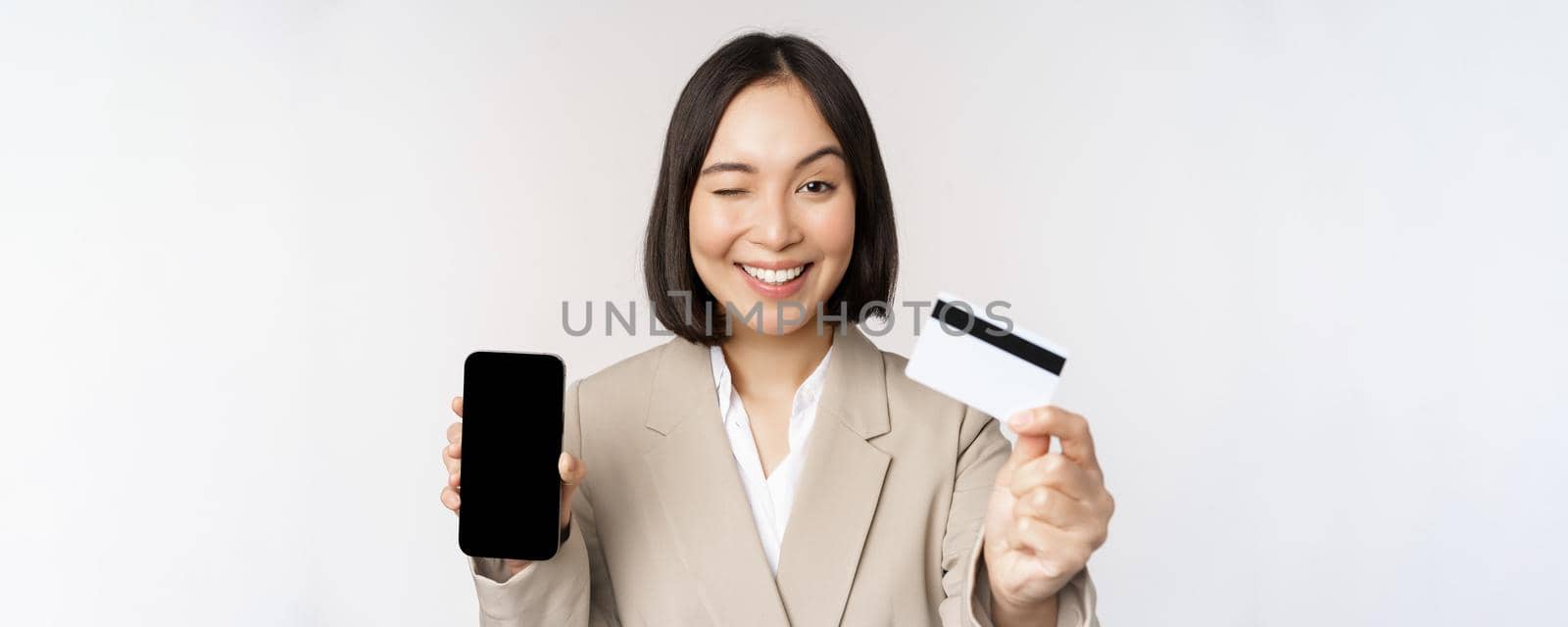 Smiling corporate woman in suit, showing mobile phone screen and app on mobile phone, smartphone screen, standing over white background.