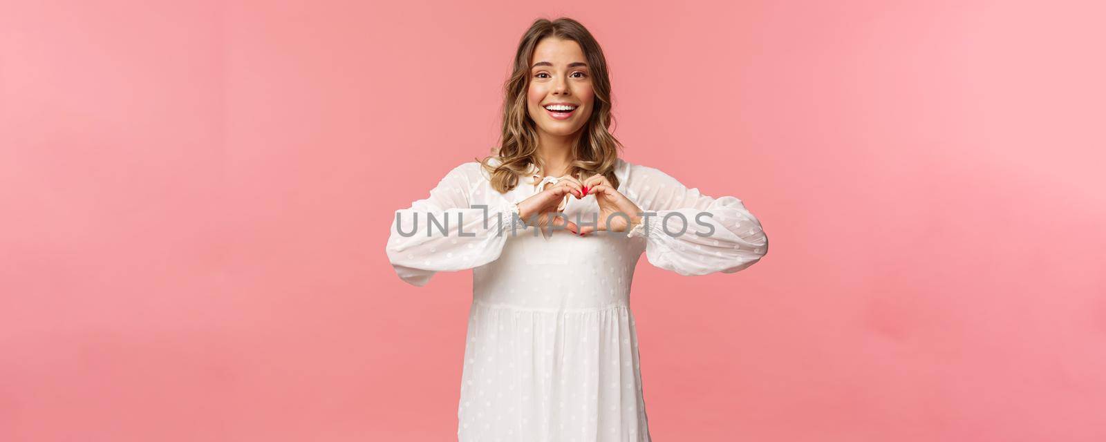 Beauty, fashion and women concept. Romantic passionate blond european female in white cute dress, make heart sign near chest and smiling at camera excited, show sympathy.