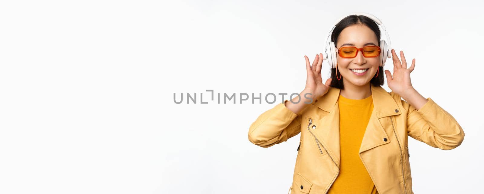 Beautiful asian girl, laughing happy, listening music in headphones, standing over white background. Copy space