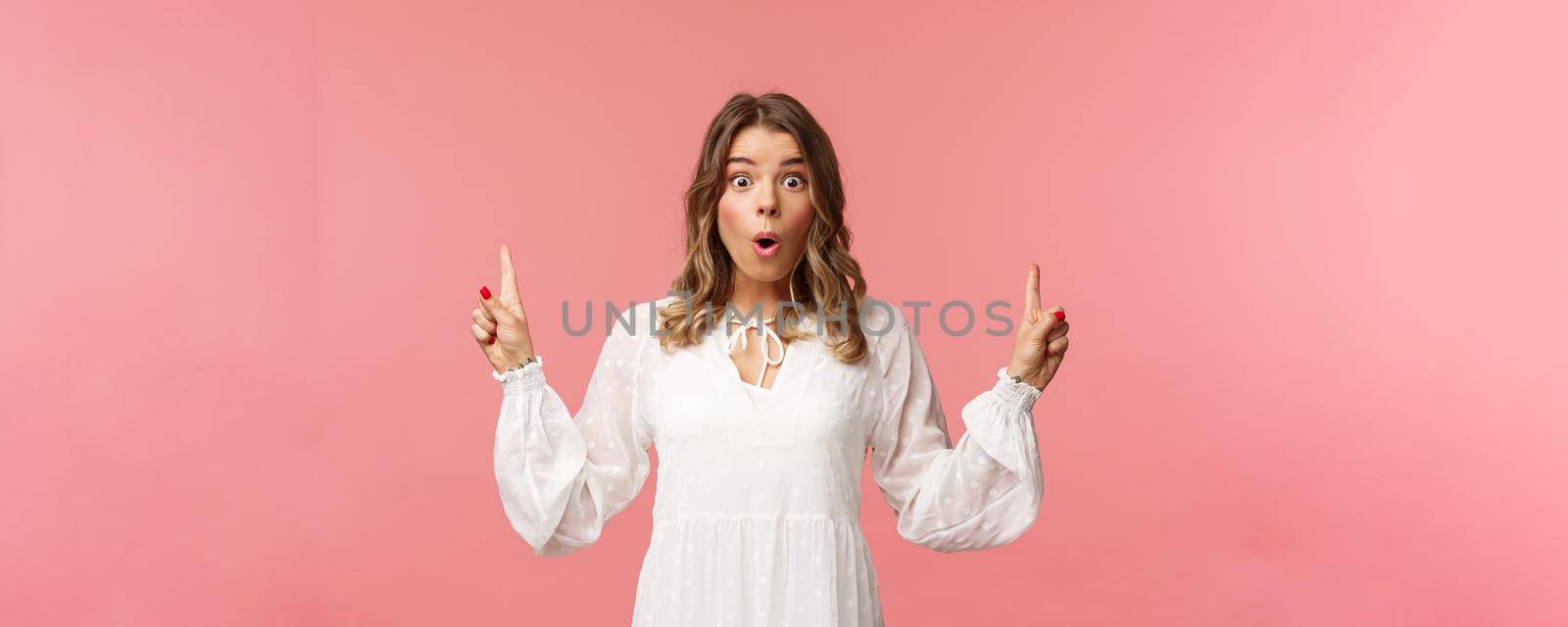 Waist-up portrait of amazed blond girl say wow in excitement, pointing fingers up, showing person top advertisement with cool special spring discount, standing pink background.