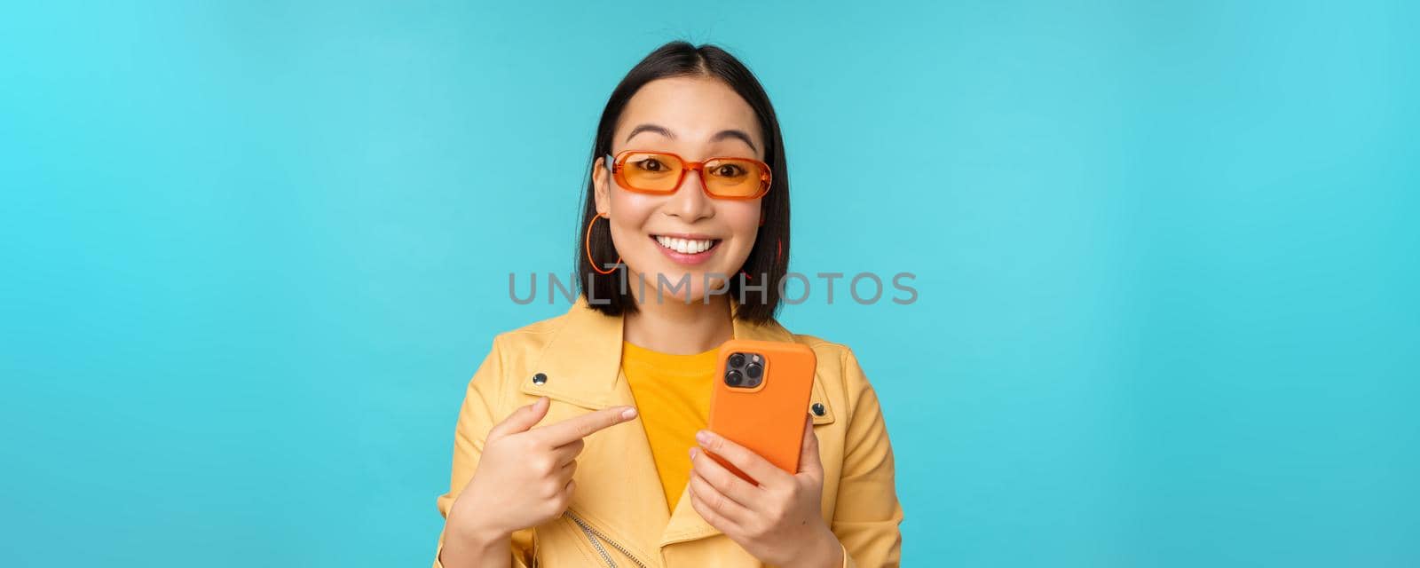 Enthusiastic asian girl in stylish sunglasses, pointing finger at smartphone, showing mobile phone, standing over blue background.