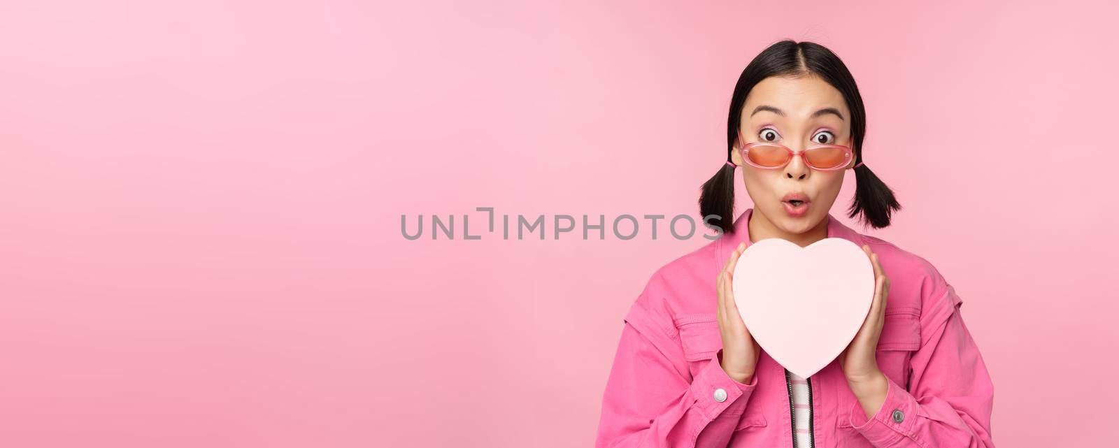 Beautiful asian girl smiling happy, showing heart gift box and looking excited at camera, standing over pink romantic background.