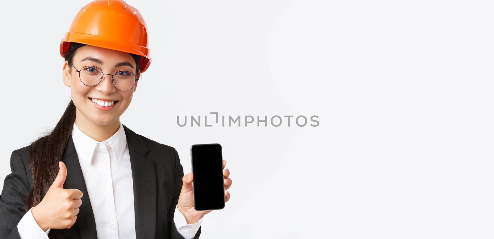 Close-up of professional smiling female engineer, construction manager in business suit and safety helmet, showing smartphone screen and thumbs-up in approval, white background by Benzoix