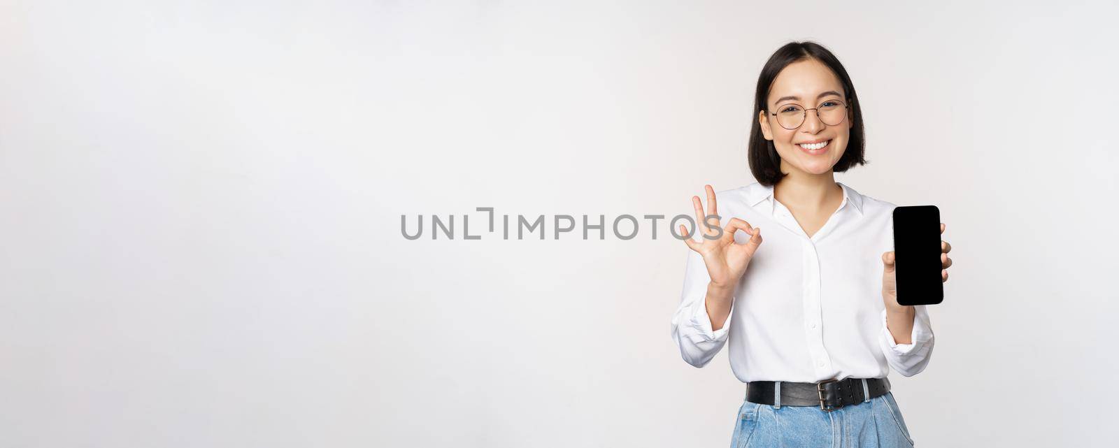 Image of asian businesswoman, showing smartphone screen, app interface and ok sign, recommending application on mobile phone, standing over white background.