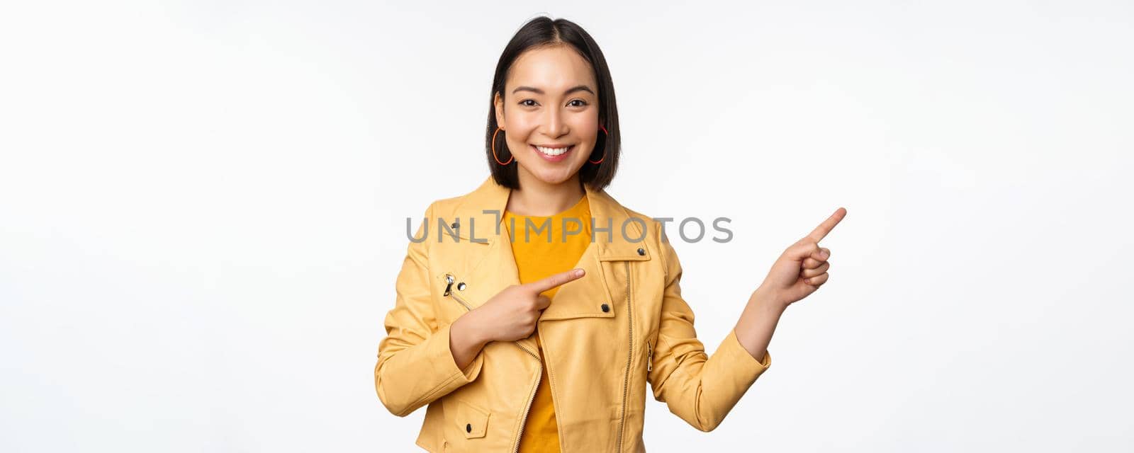 Happy asian woman smiling, pointing fingers right, inviting to check out sale, showing advertisement banner or logo, standing over white background.