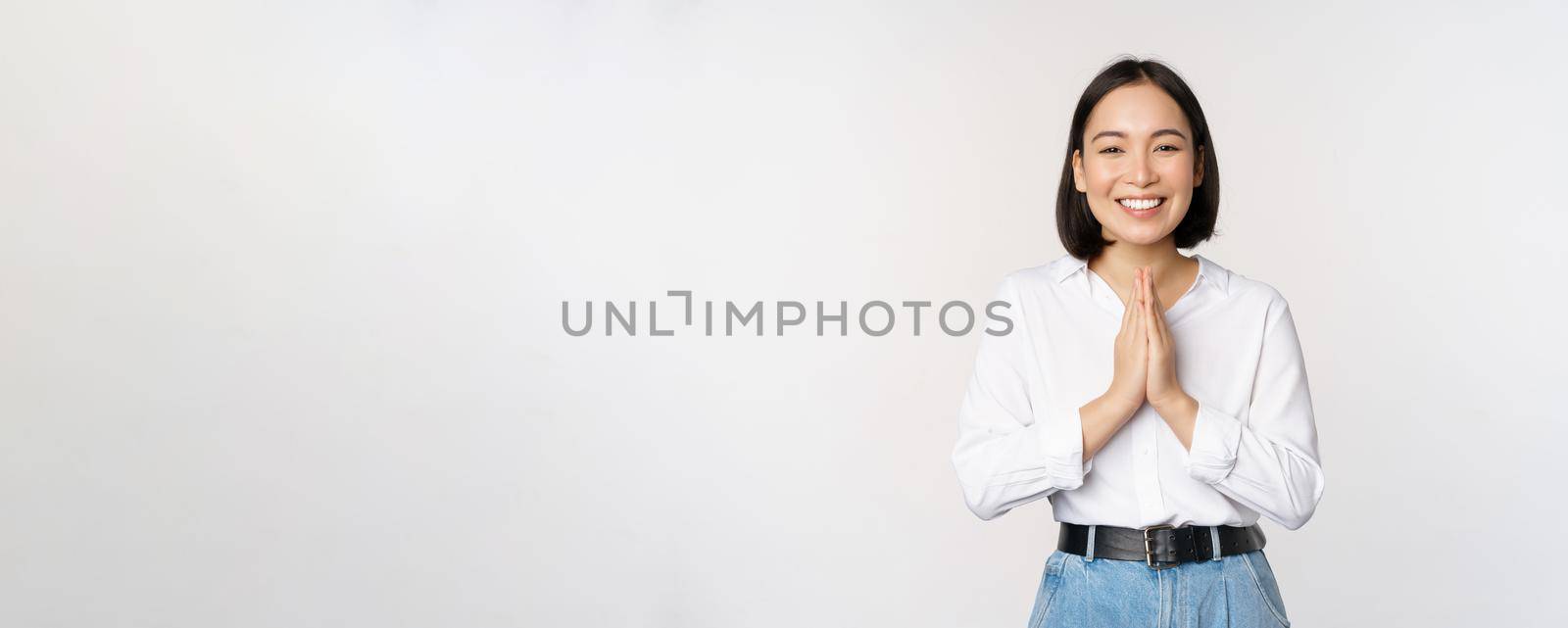 Portrait of happy asian girl laughing and smiling, showing thank you, namaste gesture, grateful for smth, standing over white background.