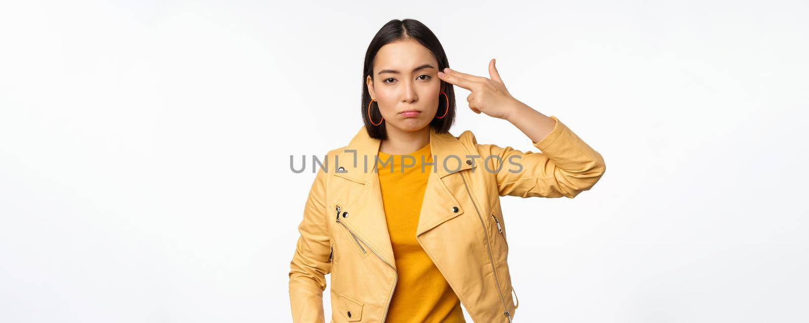 Image of annoyed and tired asian girl, making hand-gun gesture, fingers near temple, looking bored and distressed, standing against white background.