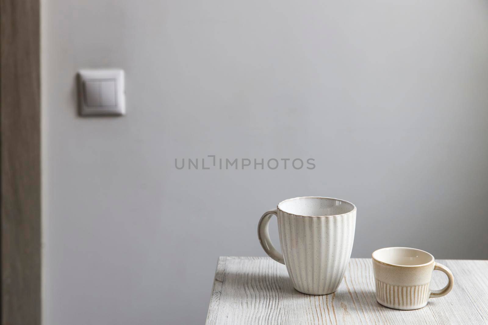 Two porcelain white cups are on the beige table. Large and small cup by elenarostunova