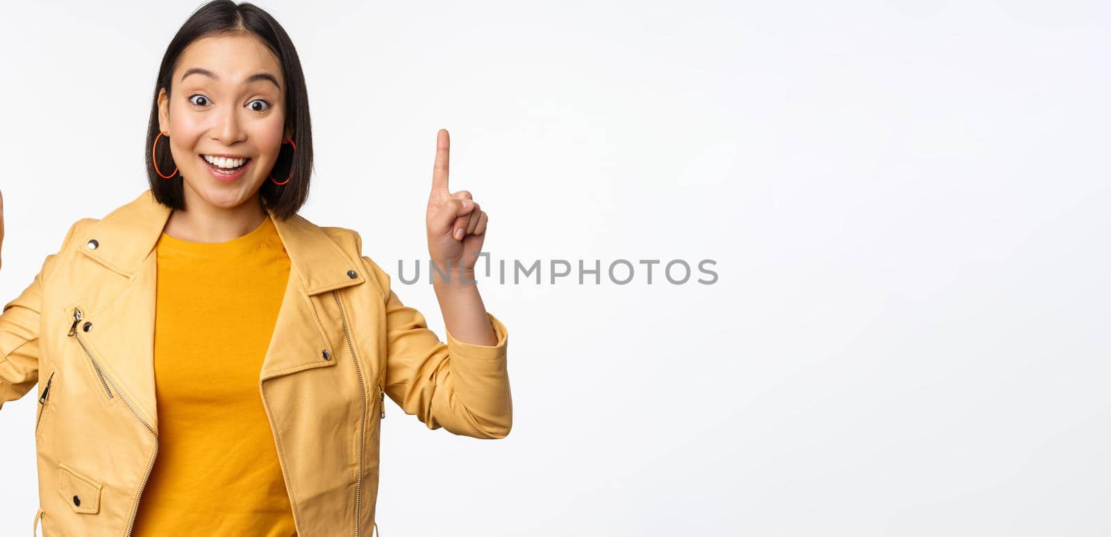 Enthusiastic asian girl pointing fingers up, showing advertisement on top, smiling happy, demonstrating promo offer or banner, standing over white background.