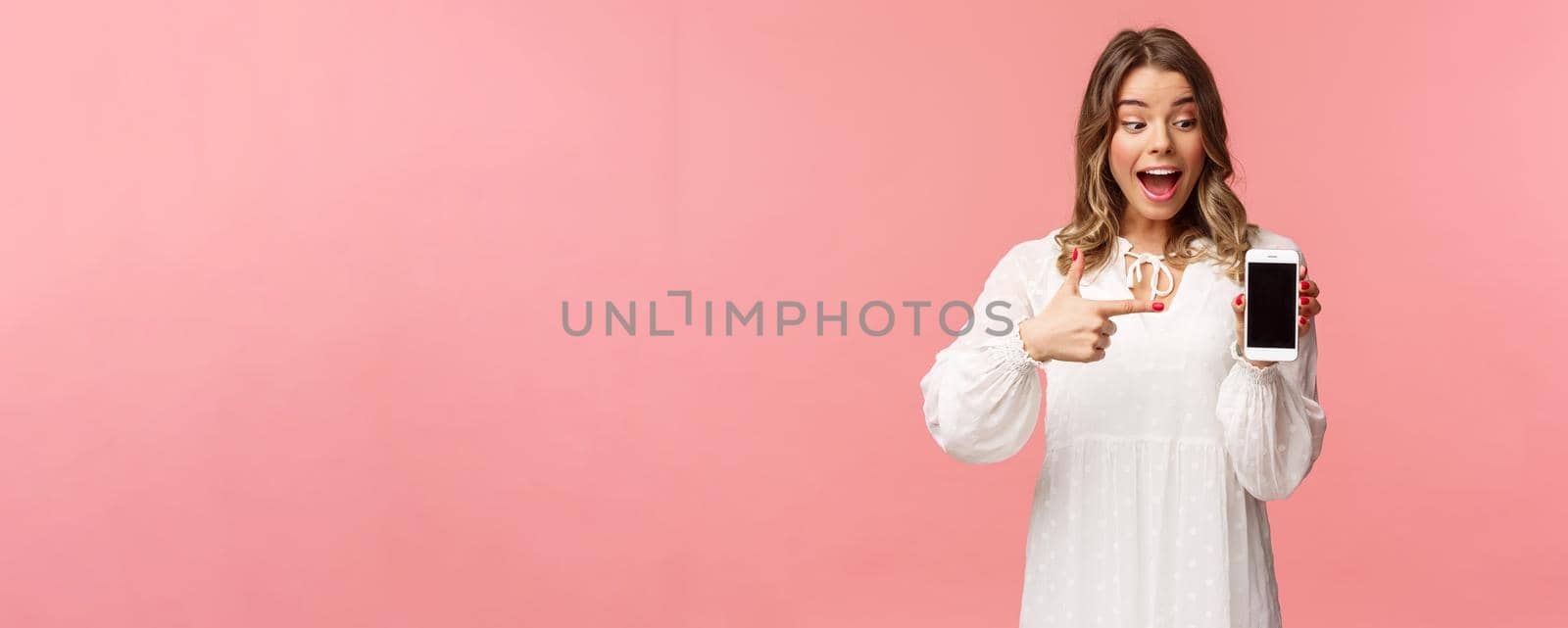 Portrait of excited and amazed, happy blond female in white dress, pointing finger at mobile phone screen, looking at smartphone with pleased and surprised smile, stand pink background.