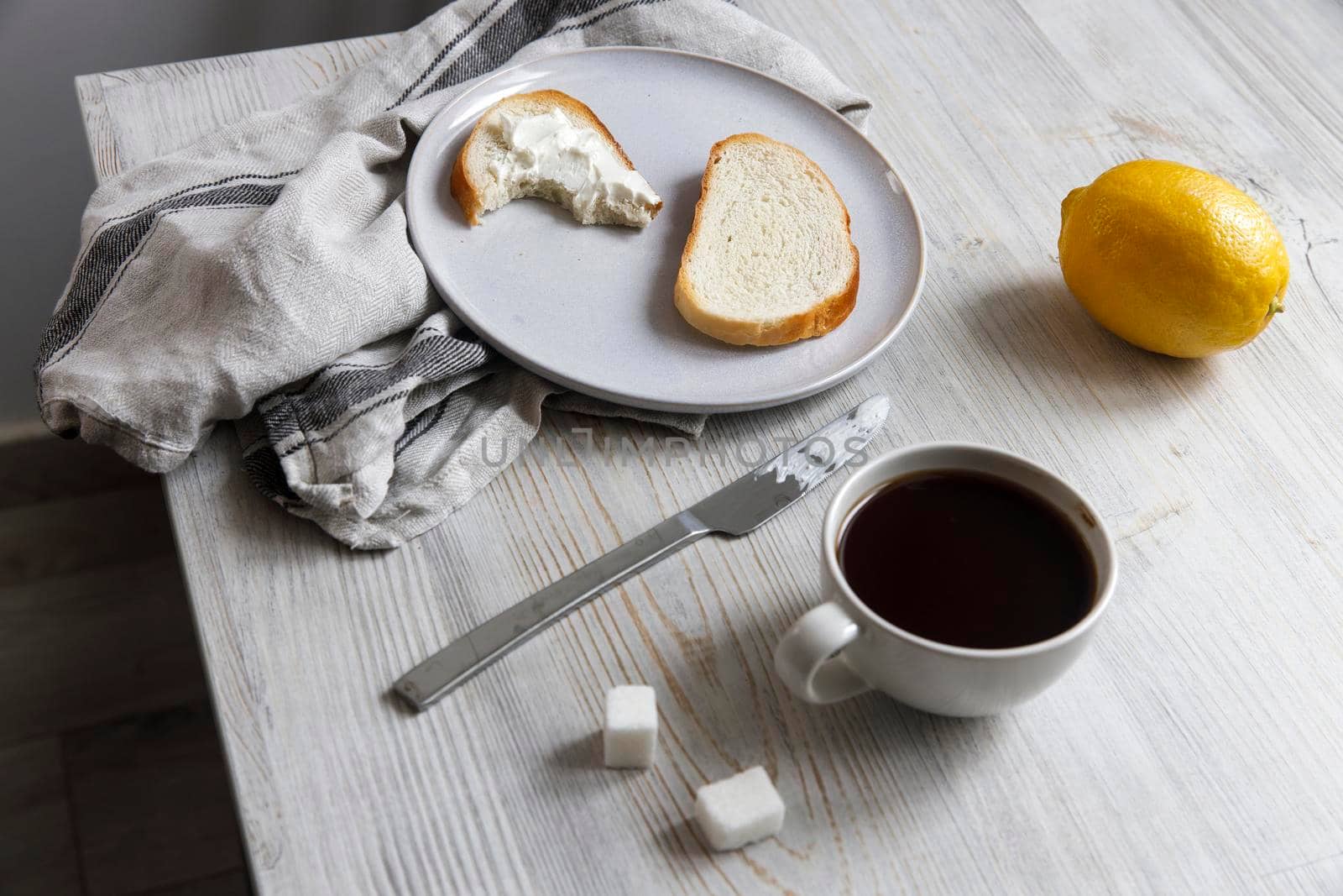 A cup of coffee, two pieces of sugar, a lemon, a piece of bread with curd cheese spread on it on a white ceramic plate with a knife on the table. by elenarostunova