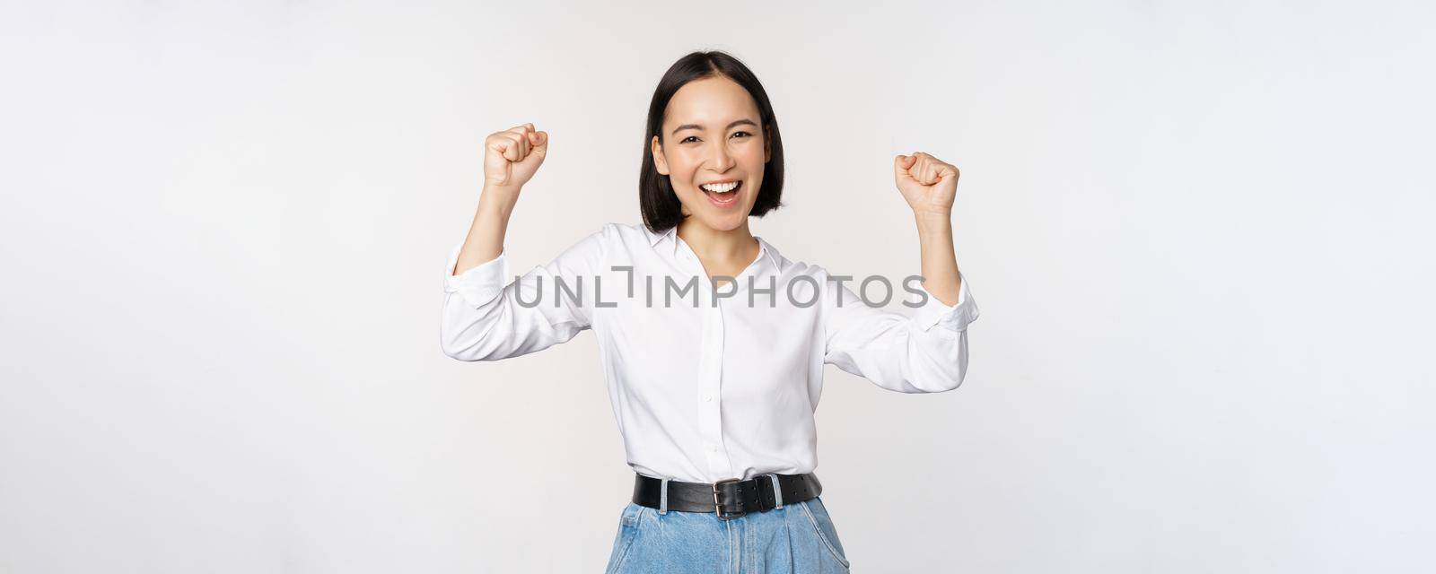 Enthusiastic asian woman rejoicing, say yes, looking happy and celebrating victory, champion dance, fist pump gesture, standing over white background.