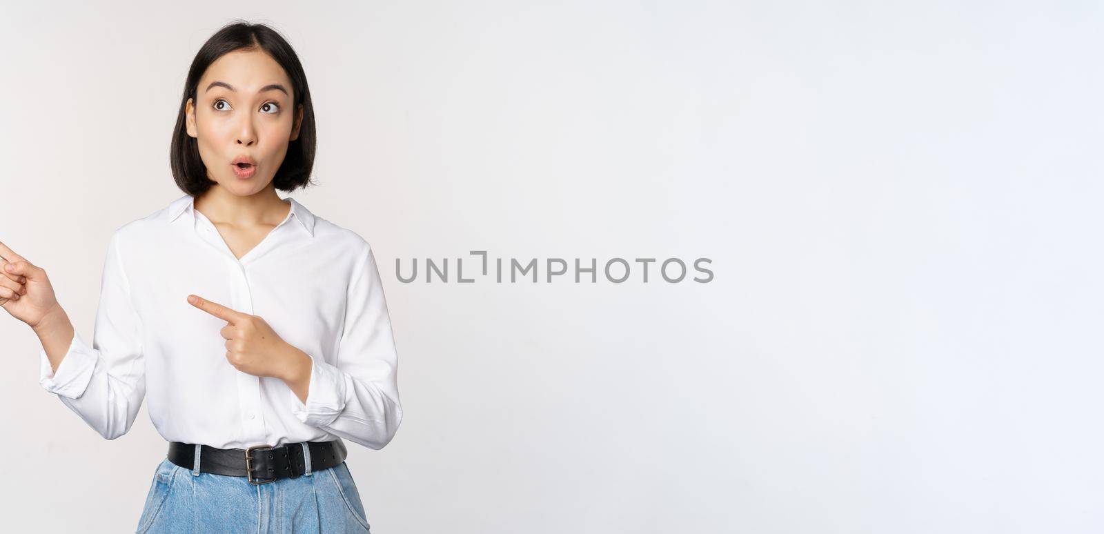 Portrait of attractive adult asian woman pointing, looking left with pleased smile, showing banner or logo aside, standing against white studio background.