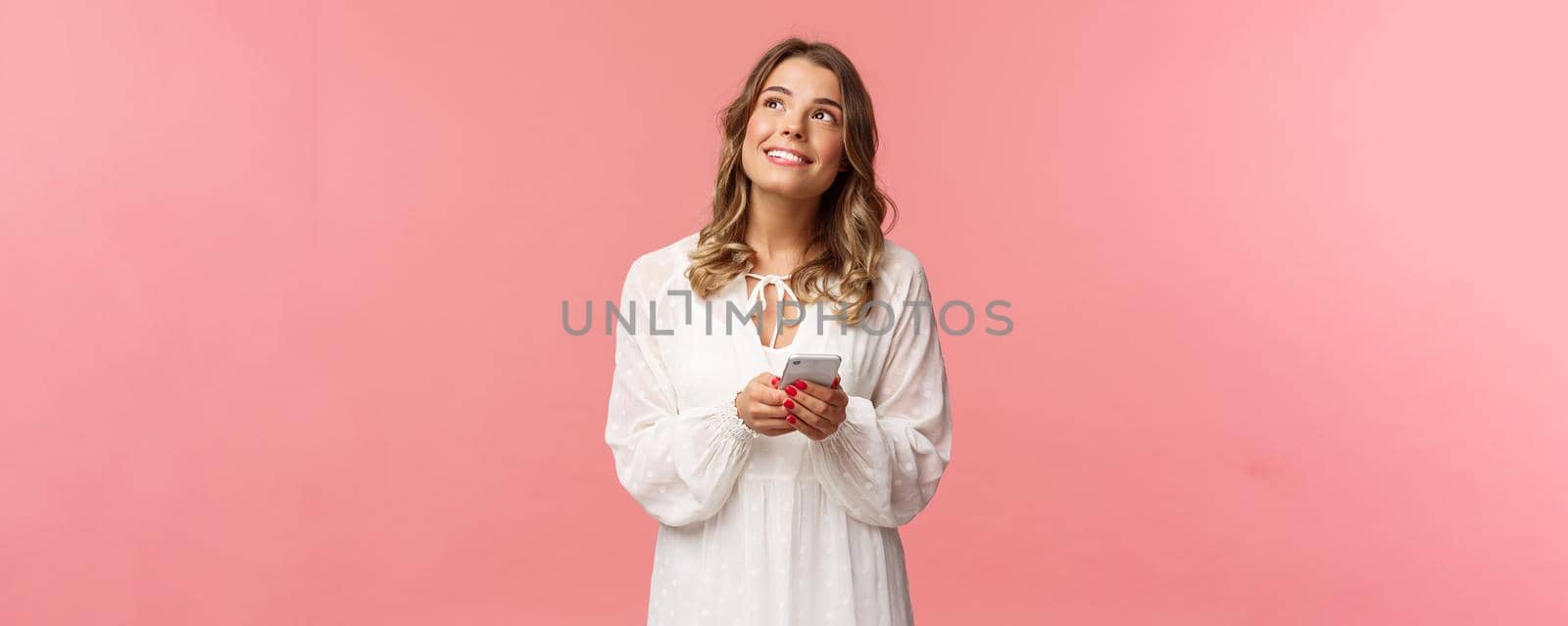 Portrait of dreamy young pretty blond girl fantasizing about something beautiful, looking up thoughtful and happy smiling, holding mobile phone, messaging, shopping online, pink background.