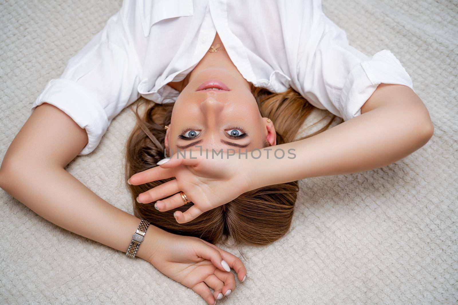 The woman lies on the bed on her back, top view. She looks straight ahead, wearing a white shirt by Matiunina