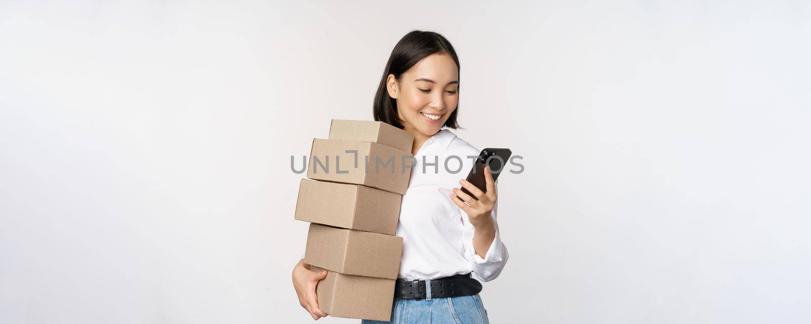 Image of young asian woman holding boxes, customer orders and looking at mobile phone, standing over white background.