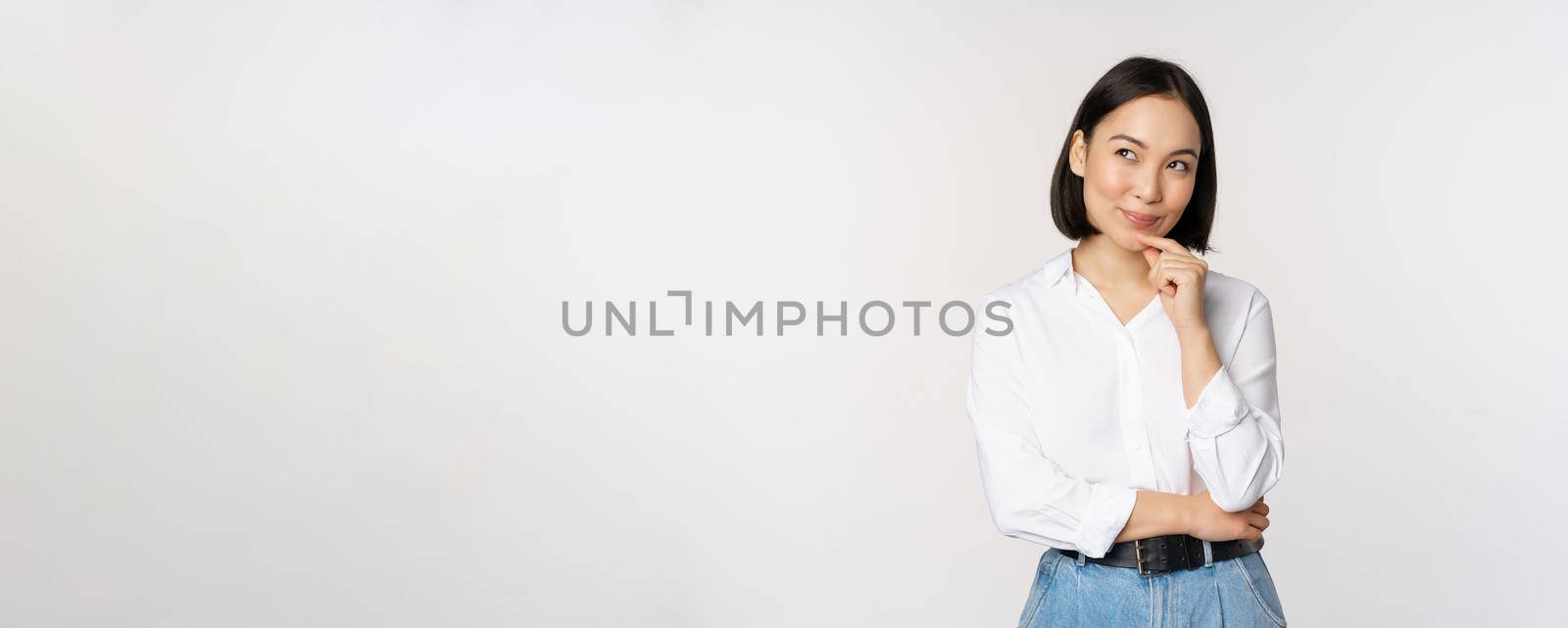 Image of thoughtful smiling woman has an idea, scheming, planning, looking aside and thinking, standing in office white blouse against studio background.