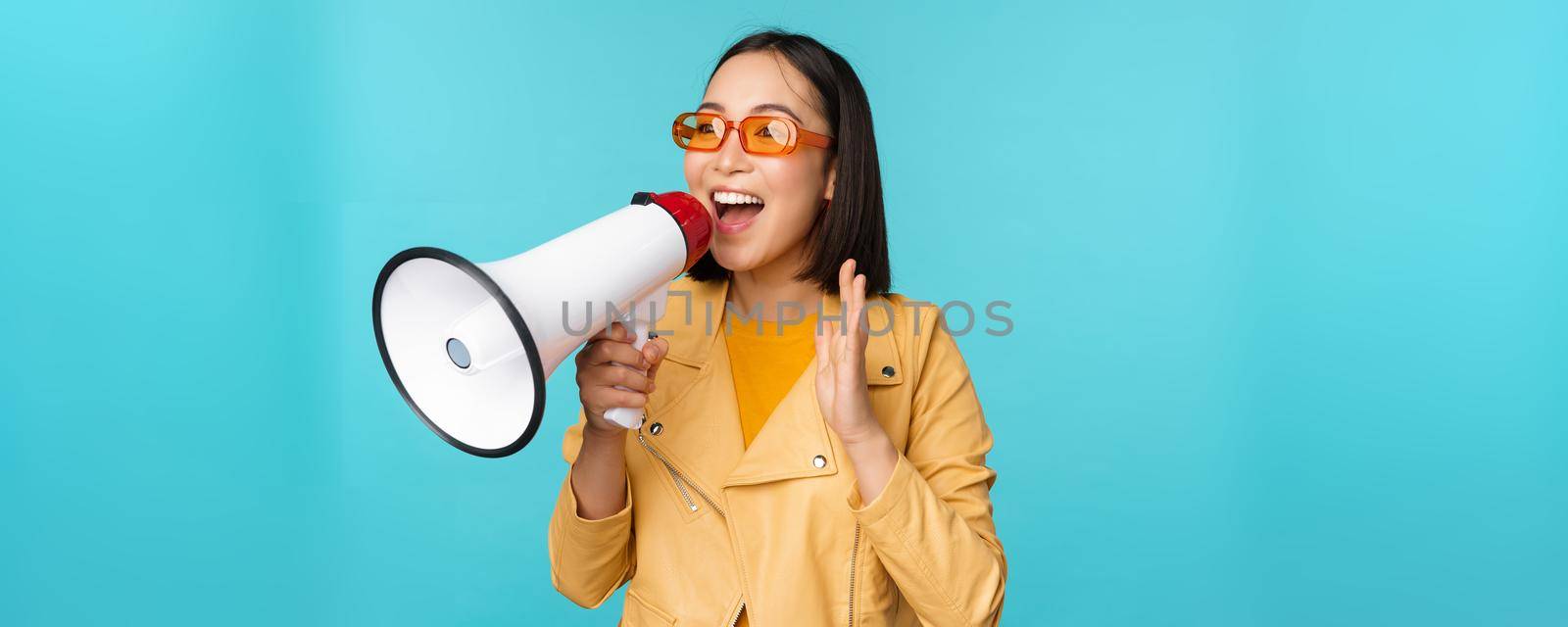 Stylish asian girl making announcement in megaphone, shouting with speakerphone and smiling, inviting people, recruiting, standing over blue background.