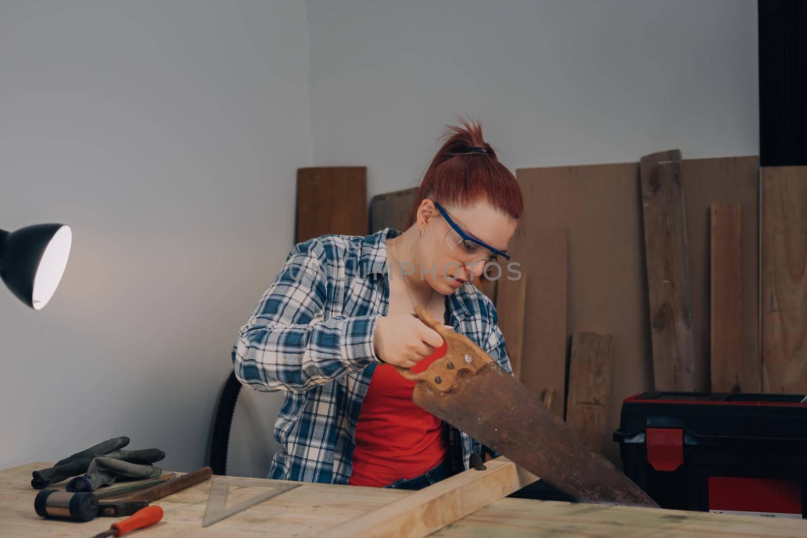 young red-haired female carpenter, concentrated and precise, working on the design of wood in a small carpentry workshop, dressed in blue checked shirt and red t-shirt. Young businesswoman sawing a wooden board in her small workshop and designing new furniture for the house. Warm light indoors, background with wooden slats. Horizontal.