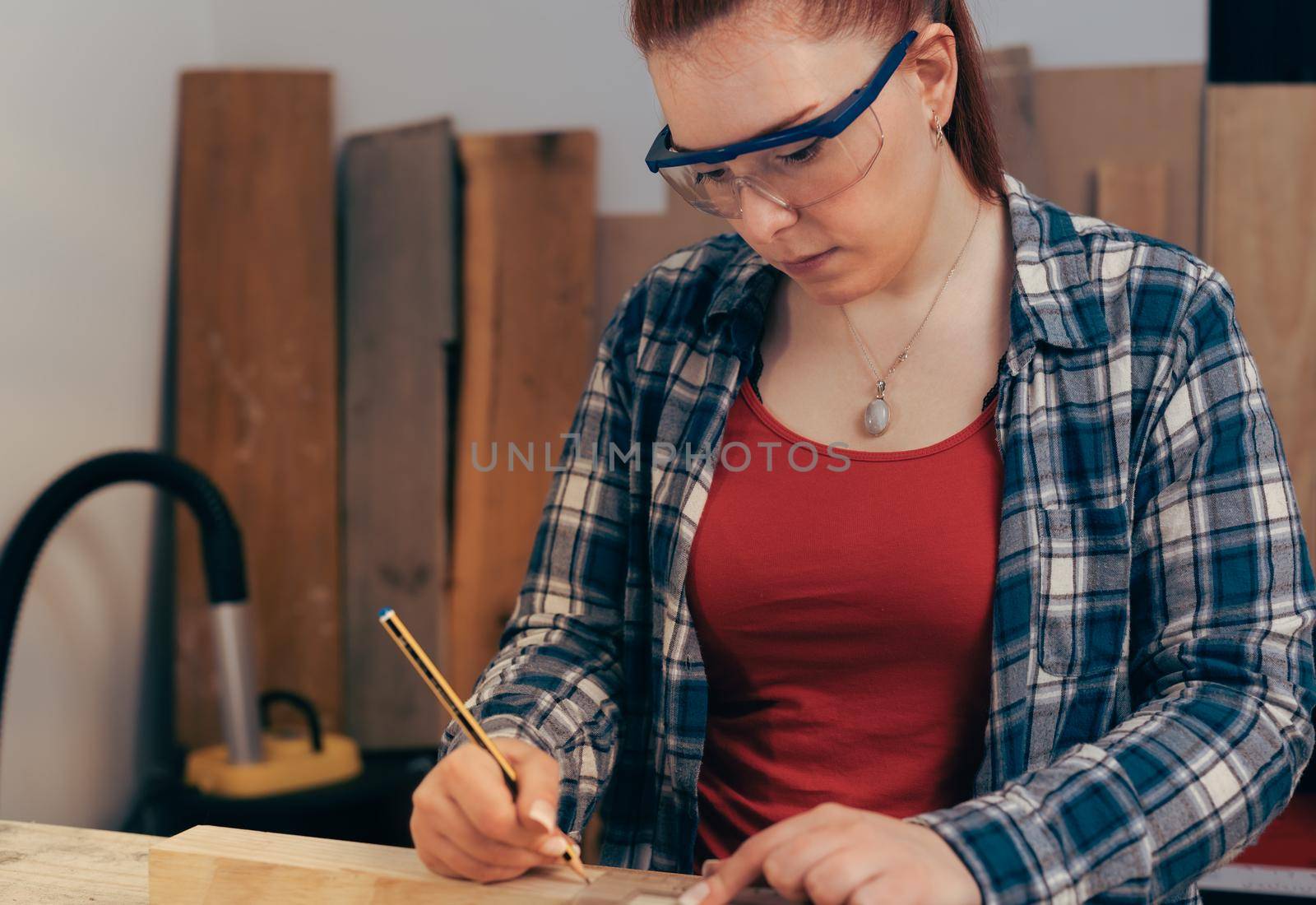 Young red haired carpenter woman, concentrated and precise, working on wood design in a small carpentry workshop, dressed in blue checked shirt and red t-shirt. Young businesswoman measuring a wooden board in her small workshop and designing new furniture for the house. Warm light indoors, background with wooden slats. Horizontal.