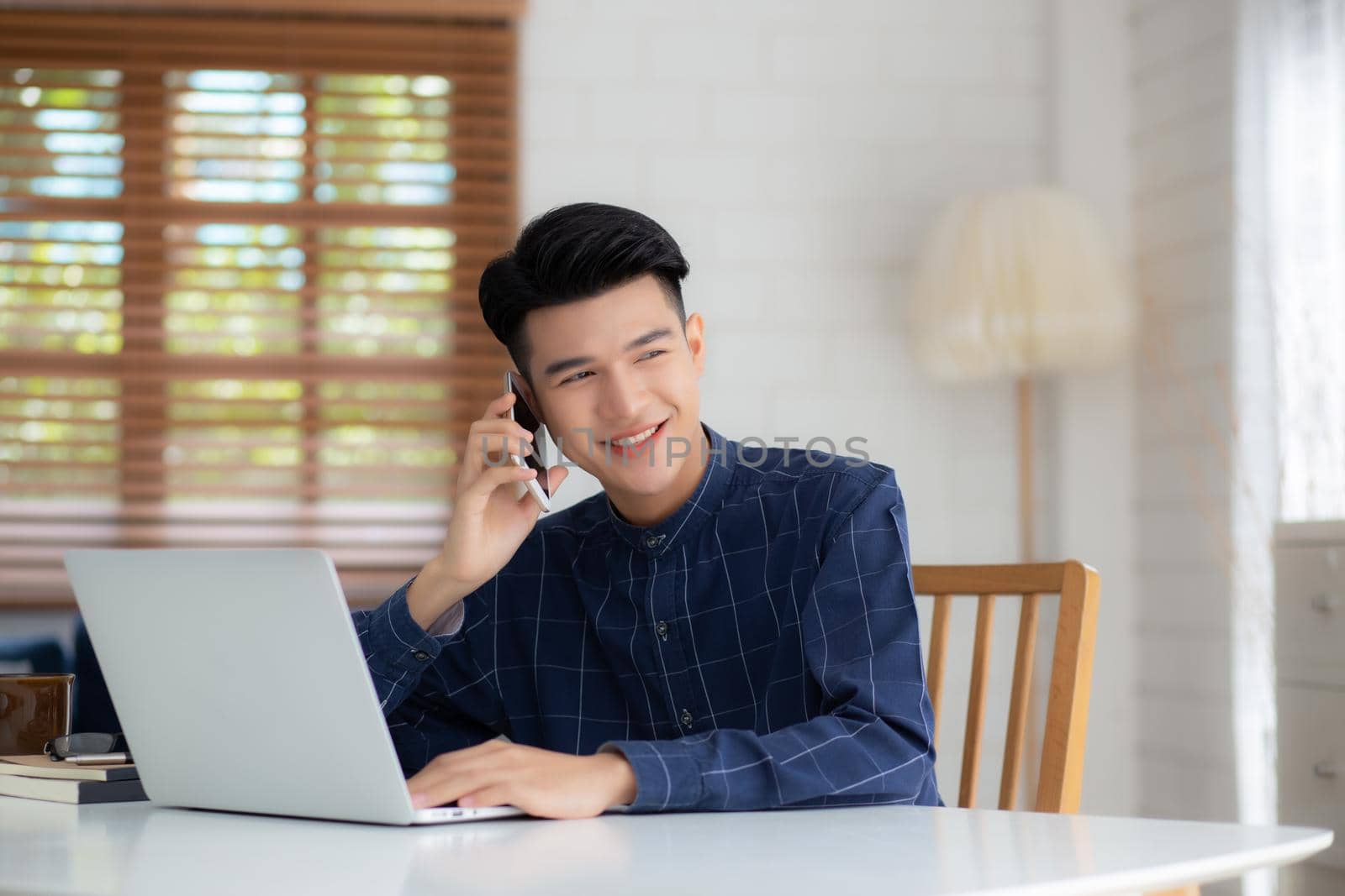 Young asian man talking phone and work from home with laptop computer, freelance using notebook to internet online and speak on smartphone, domestic life, business and communication concept.