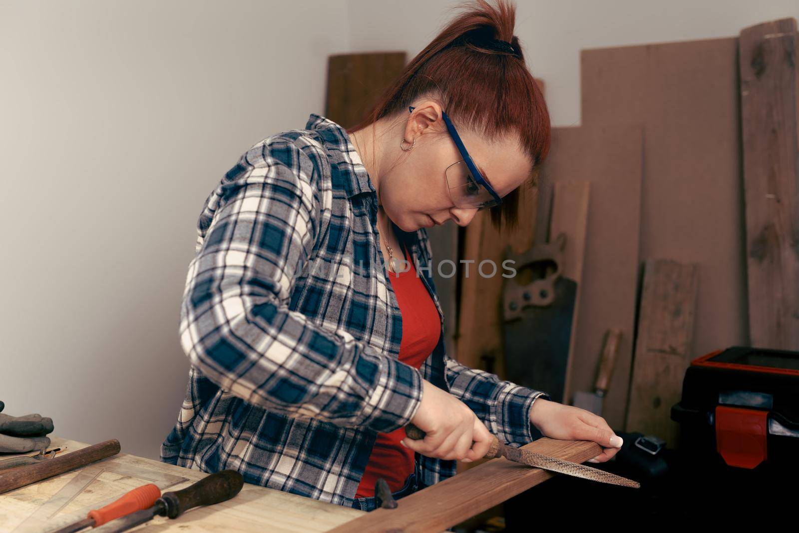 young red-haired carpenter woman, concentrated and precise, working on the design of wood in a small carpentry workshop, dressed in a blue checked shirt and a red t-shirt. Carpenter woman holding a nail in a wooden board, in her small carpentry business. Warm light indoors, background with wooden slats. Horizontal.